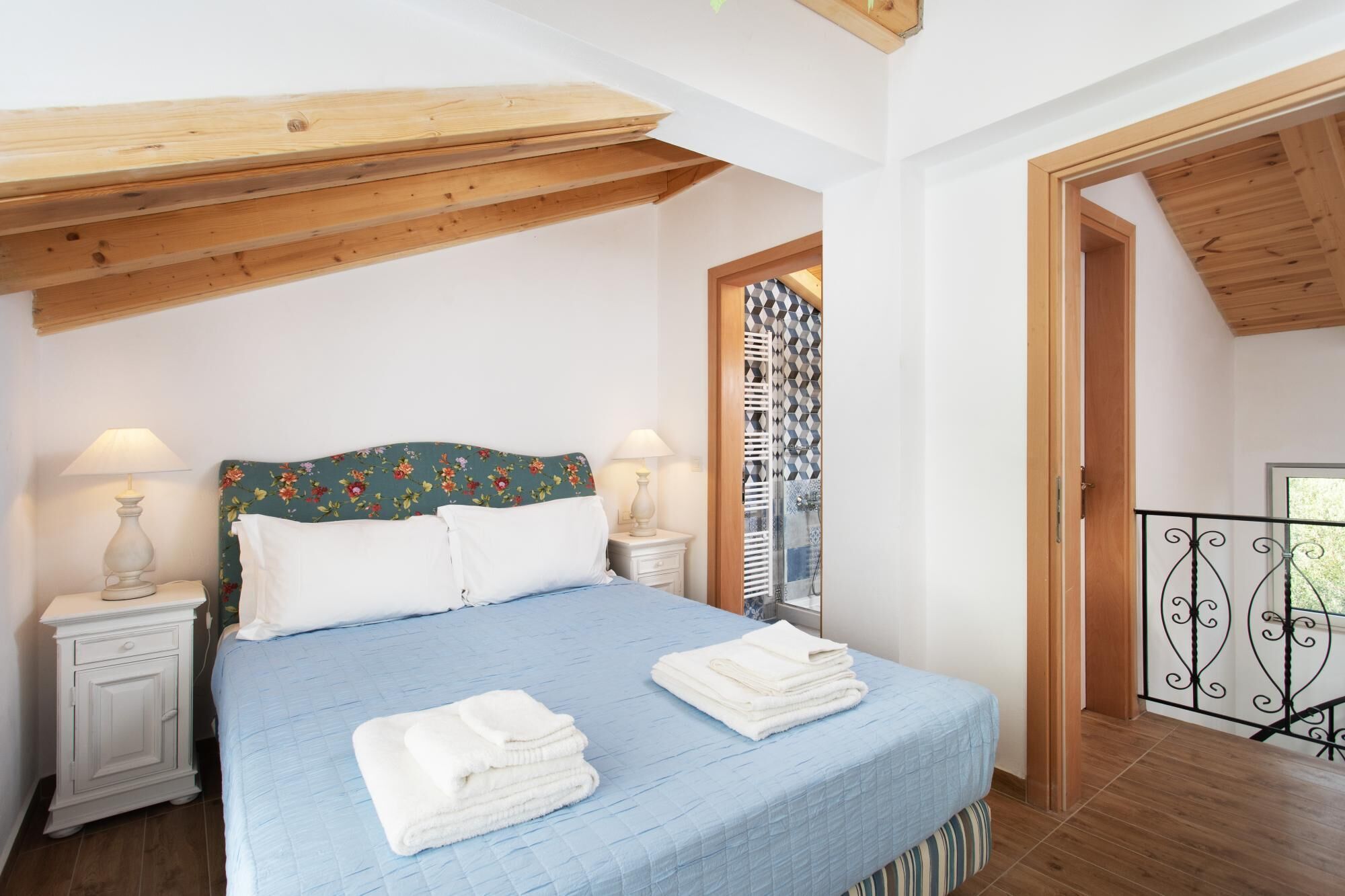 A bedroom with a wooden ceiling, a bed with a blue quilt, white pillows, and two bedside lamps. There are folded towels on the bed and a small bedside table.