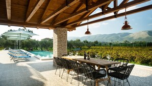 A covered outdoor area with a pool, dining table, chairs, and a view of vineyards and mountains. - Aneli Villas - Villa Alexandra (Zakynthos)