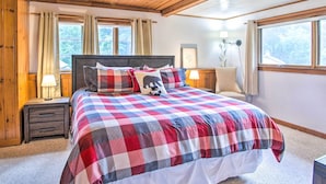A bedroom with a large bed, wooden headboard, two bedside tables with lamps, a window with curtains, and a view of trees outside. - Beautiful Carriage House in Downtown Jackson! (Jackson)