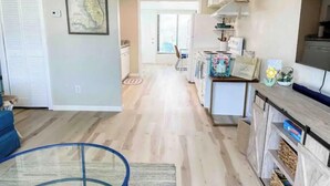 A spacious living room with a blue sofa, a glass coffee table, and a kitchen area with white cabinets and a TV. - Cute Beach House half a block from the beach 🏝 (Ormond Beach)