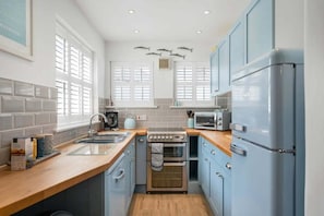 A kitchen with a stainless steel stove, a microwave, and a refrigerator. There are windows with shutters and a tiled backsplash. - Beautiful Cottage Minutes from Mevagissey Harbour (Saint Austell)