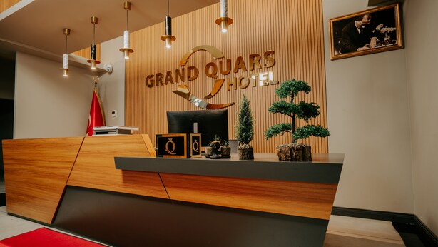 Taulell de recepció a l'Hotel Grand Quars, amb un fons de fusta, un retrat emmarcat i una catifa vermella.