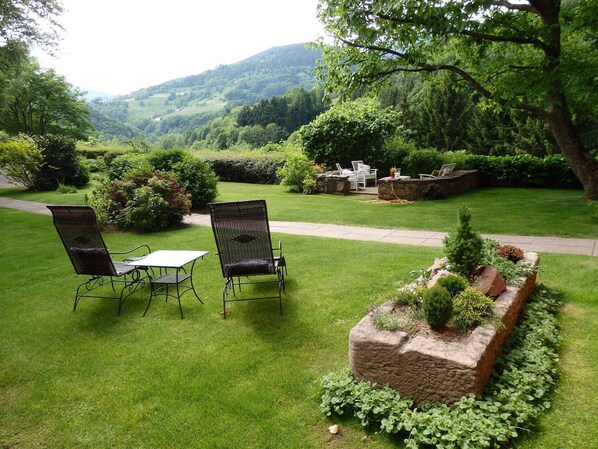 Un jardin bien entretenu, agrémenté de deux chaises en métal et d’une table, entouré d’une végétation luxuriante et d’un jardinière en pierre.