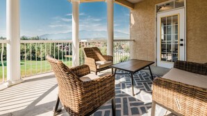 A covered patio with wicker furniture, a table, and a view of a green landscape and mountains. - Riverside Twin `A` | Balcony | King Bed | Spacious (Fairmont Hot Springs)