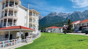 Un hotel de varios pisos con balcones, un césped y montañas al fondo.