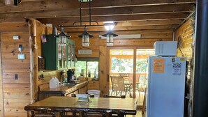 Interior rústico de cabana com mesa de jantar de madeira, cadeiras, geladeira e área de cozinha com pia e armários.