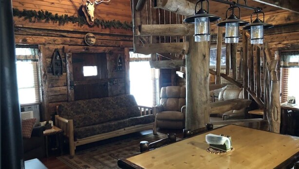 Interior rústico de cabana com mesa de jantar de madeira, sofá de couro e escada.