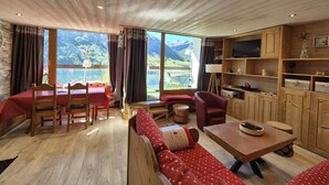 A cozy living room with a wooden ceiling, a large window with a mountain view, a dining area with a red table and chairs, and a built-in TV shelf. - 150m from resort center, 3rd floor, view lake, balcony, tv, ski locker, 40m², Tignes (Tignes)