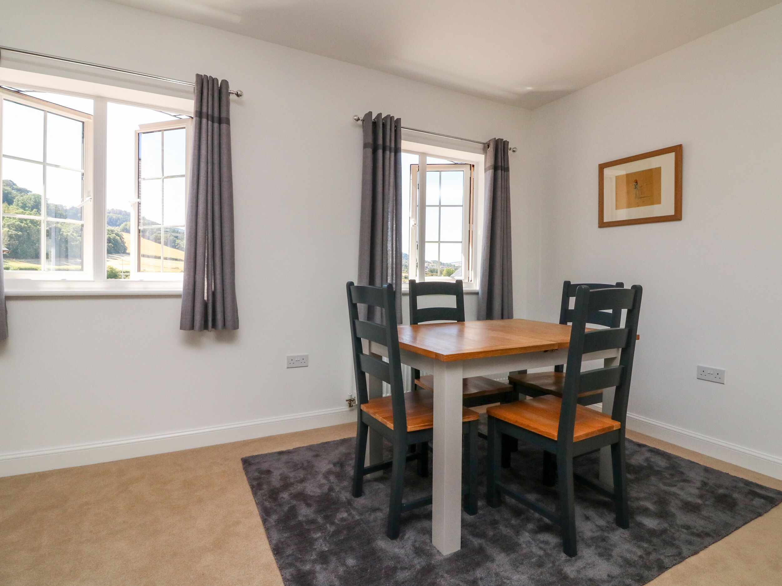 A dining area with a wooden table and four chairs, a window with curtains, and a framed picture on the wall.