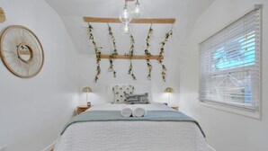 A bedroom with a bed, wooden headboard, hanging floral decorations, bedside lamps, and a window with blinds. - Sheridan Wildflower! Downtown Family Fun w/Firepit (Colorado Springs)