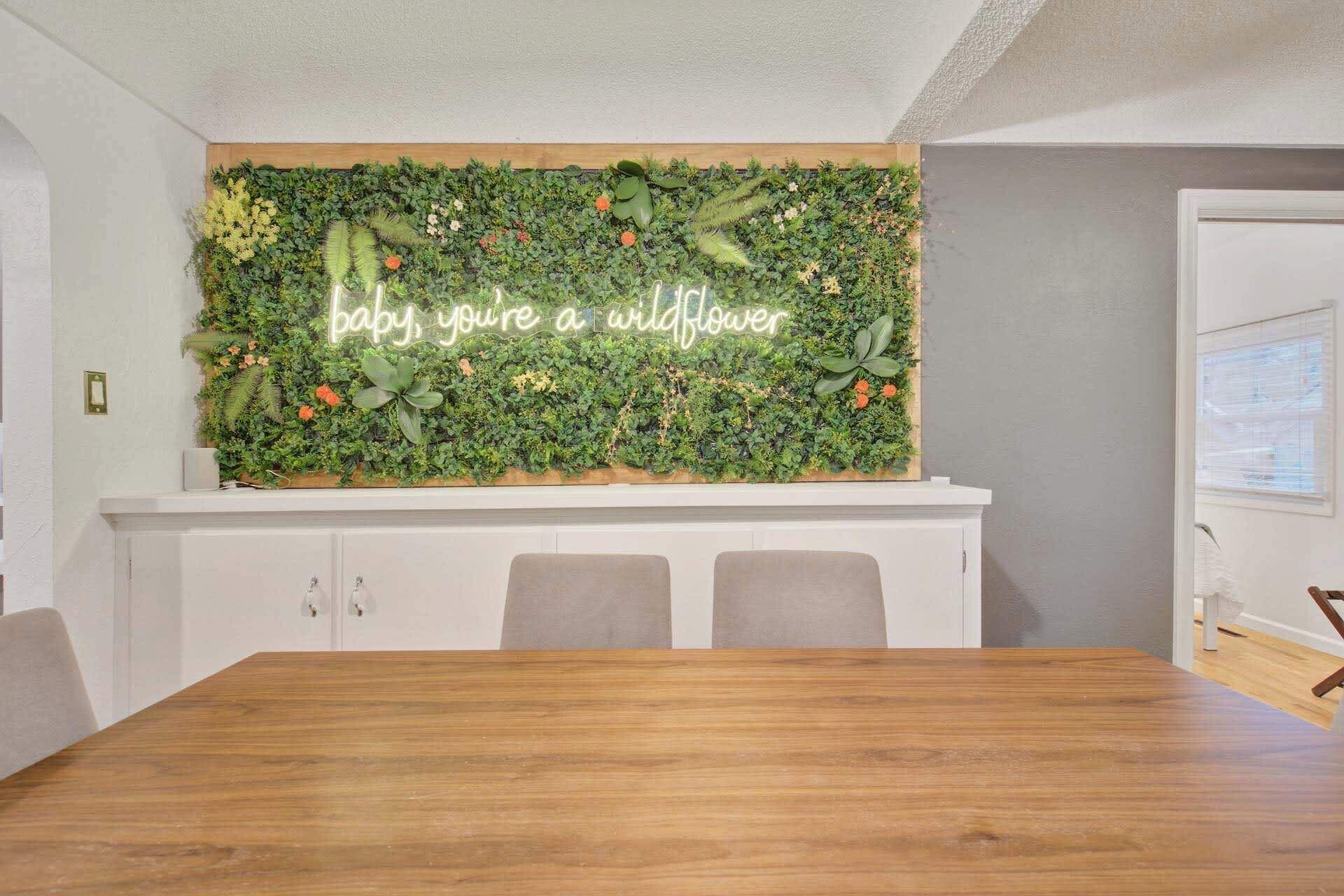 Ein Zimmer mit einem Holztisch, einem weißen Schrank und einer Wand, die mit einem grünen Wandbehang und einem Neonzeichen mit dem Schriftzug 'baby, you're a wildflower' verziert ist.
