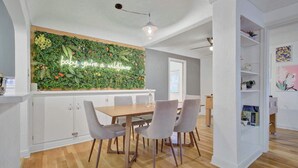 A dining area with a wooden table and grey chairs, a white cabinet, and a wall-mounted green plant wall with a neon sign. - Sheridan Wildflower! Downtown Family Fun w/Firepit (Colorado Springs)