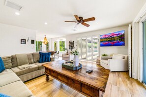 A spacious living room with a wooden coffee table, a ceiling fan, and a large TV mounted on the wall. - BAREFOOT VOLLEYBALL BUNGALOW-1ACRE (Bermuda Dunes)