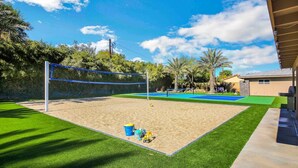 Un terrain de volleyball sablonneux avec un filet, entouré de gazon synthétique vert et de palmiers.