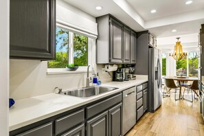 A modern kitchen with a large window, stainless steel appliances, and a dining area with a chandelier. - BAREFOOT VOLLEYBALL BUNGALOW-1ACRE (Bermuda Dunes)