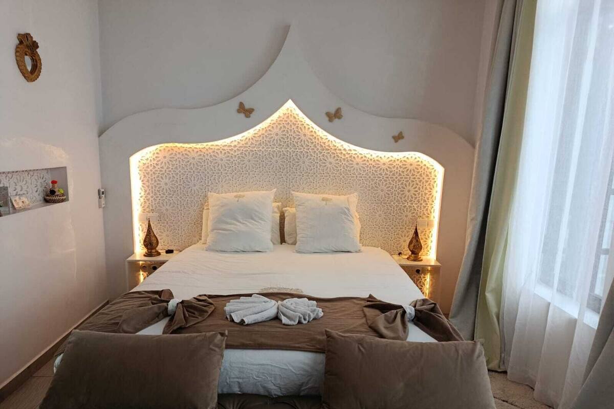 Un lit bien fait, doté d’une tête de lit capitonnée, une table de chevet avec une lampe et un élément décoratif mural.