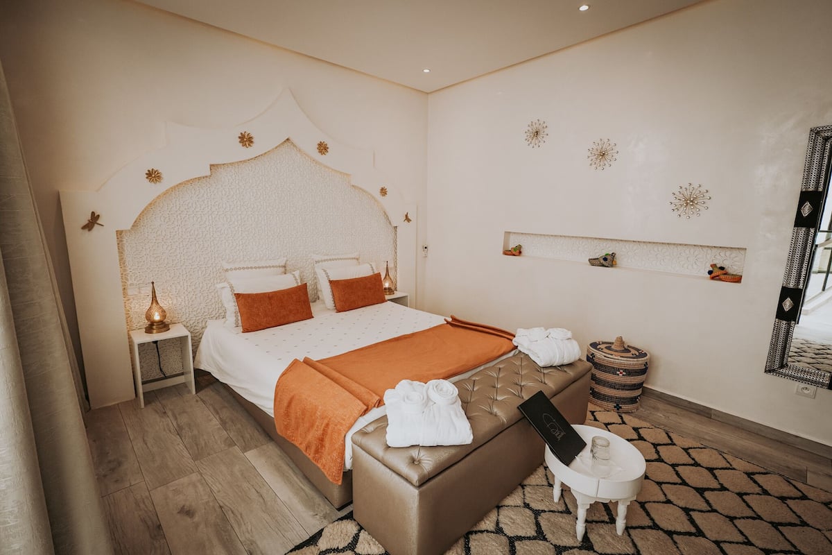 Une chambre à coucher bien aménagée, avec un grand lit, une table de chevet avec une lampe, une petite table ronde sur laquelle repose un livre, et une étagère décorative fixée au mur.