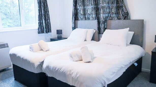 Una cama doble con sábanas blancas y dos almohadas, una mesita de noche con lámpara y una ventana con cortinas.