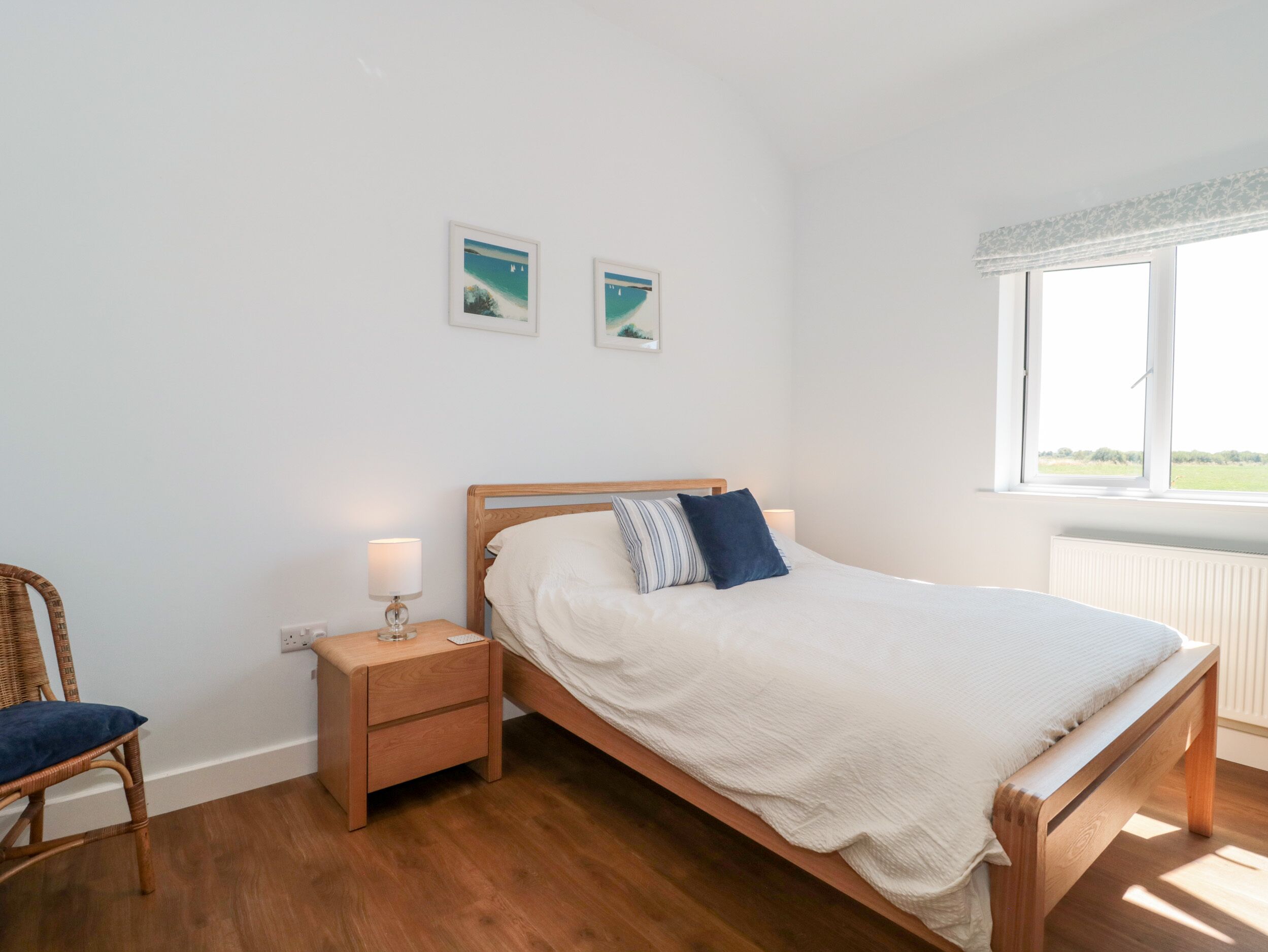 Un dormitorio con una cama de madera, una mesita de noche con lámpara, una silla de mimbre y dos cuadros en la pared.