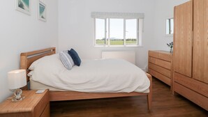 A bedroom with a wooden bed, a nightstand with a lamp, a dresser, and a window with a view of the countryside. - Robins Corner (Weston-super-Mare)