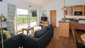 A spacious living area with a sofa, a television, and a dining table. There is a kitchen with wooden cabinets and a microwave. The room has a large sliding glass door leading to a patio with a view of the outdoors. - Robins Corner (Weston-super-Mare)