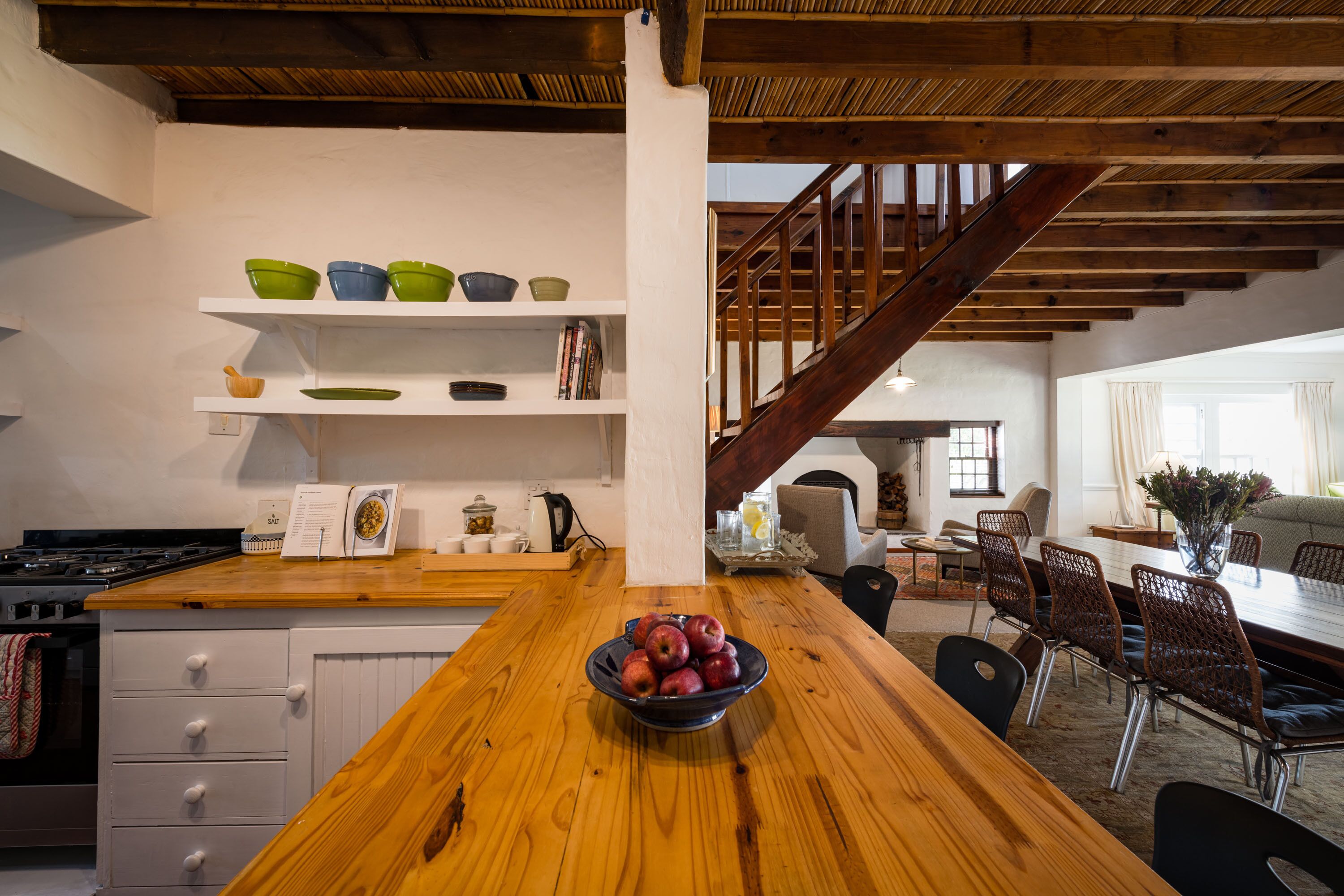 Une cuisine avec une table en bois, un saladier rempli de pommes et un escalier menant à un coin repas équipé de chaises et d’une table.