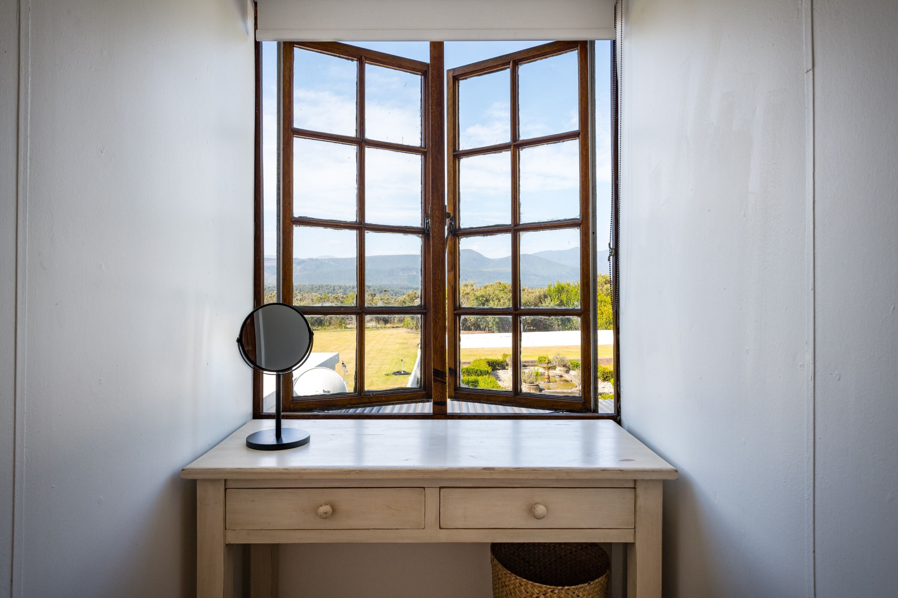 A corner desk with a mirror and a view of a landscape through an open window.