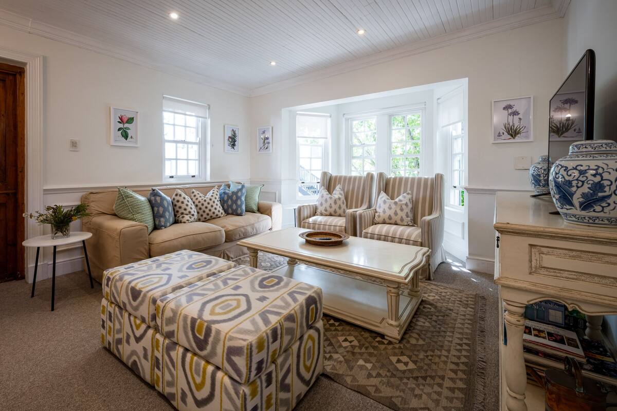 A spacious living room with a beige sofa, patterned ottoman, and a white coffee table. There are multiple windows allowing natural light, and a decorative vase on a side table.