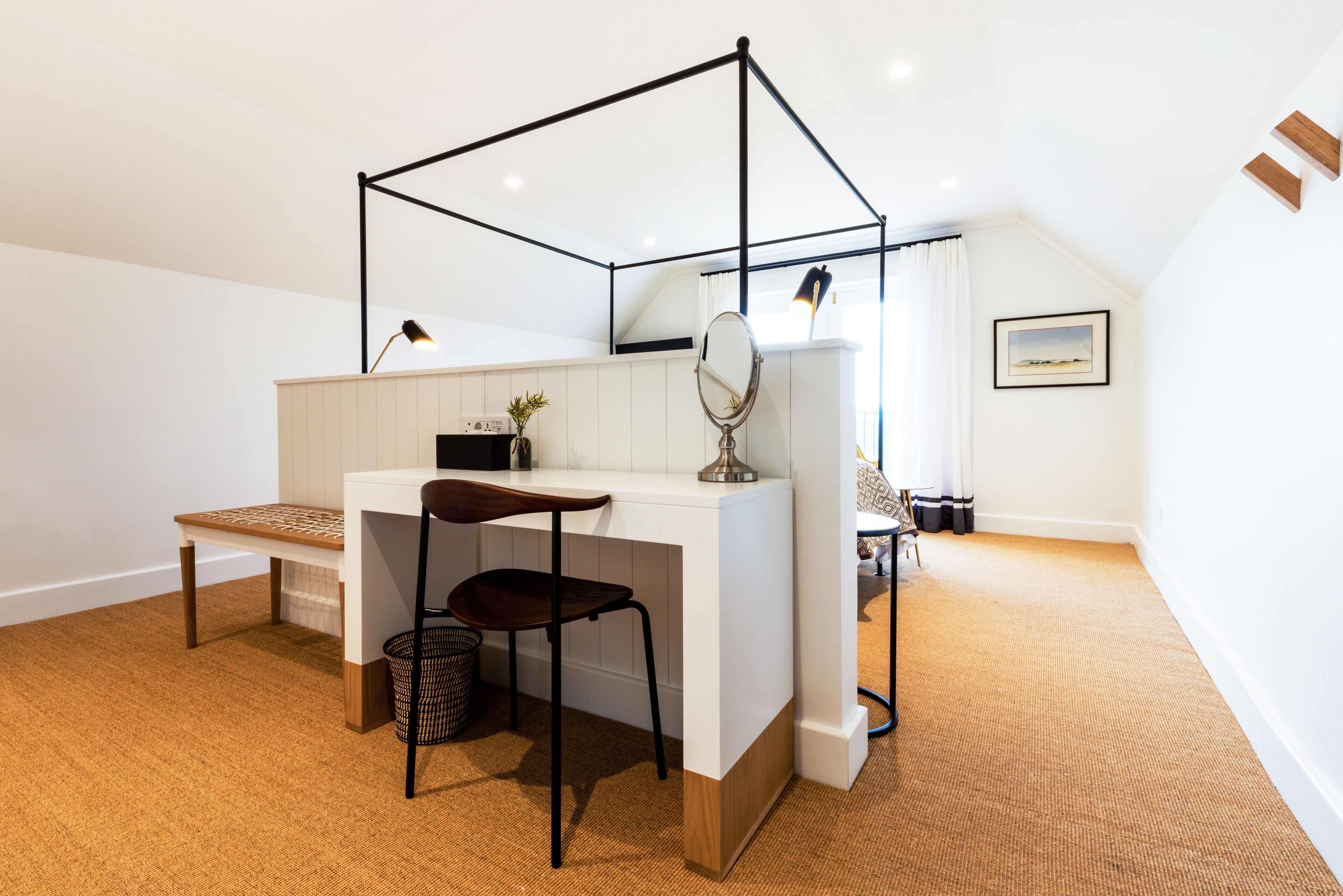Een slaapkamer met een wit bureau, een stoel, een kleine plant, een spiegel en een bed met een wit hoofdeinde.