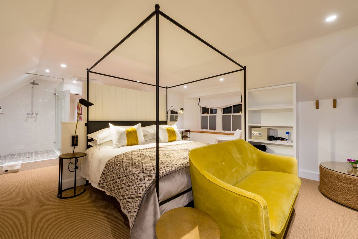 A modern bedroom with a four-poster bed, a yellow armchair, and a glass shower enclosure.