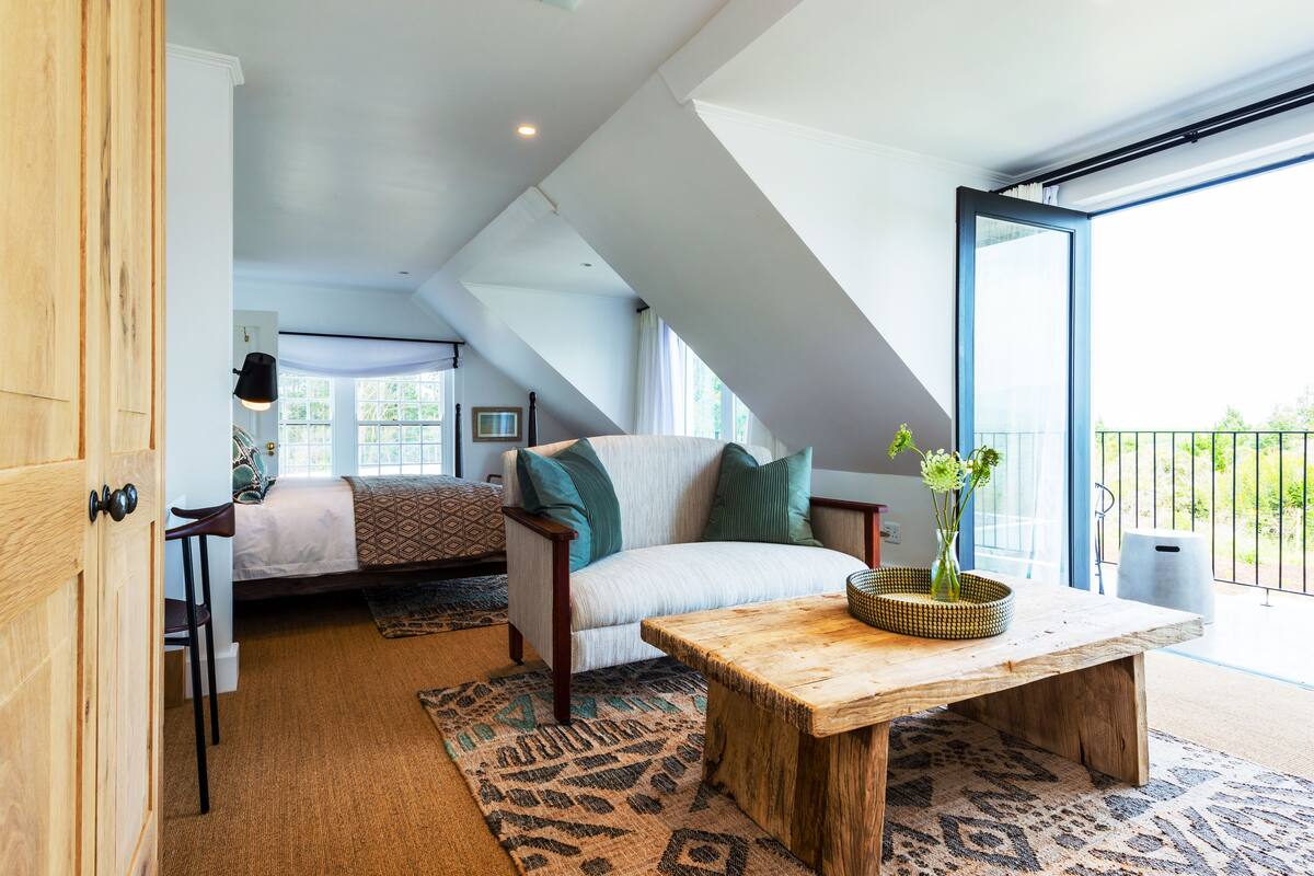 A spacious loft bedroom with a large window, a wooden table, and a cozy seating area.