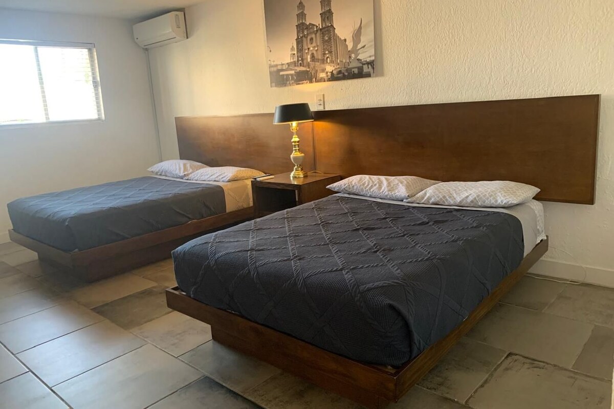 Habitación de hotel con dos camas individuales, una mesita de noche con lámpara, un cuadro grande en la pared y una ventana con persianas.