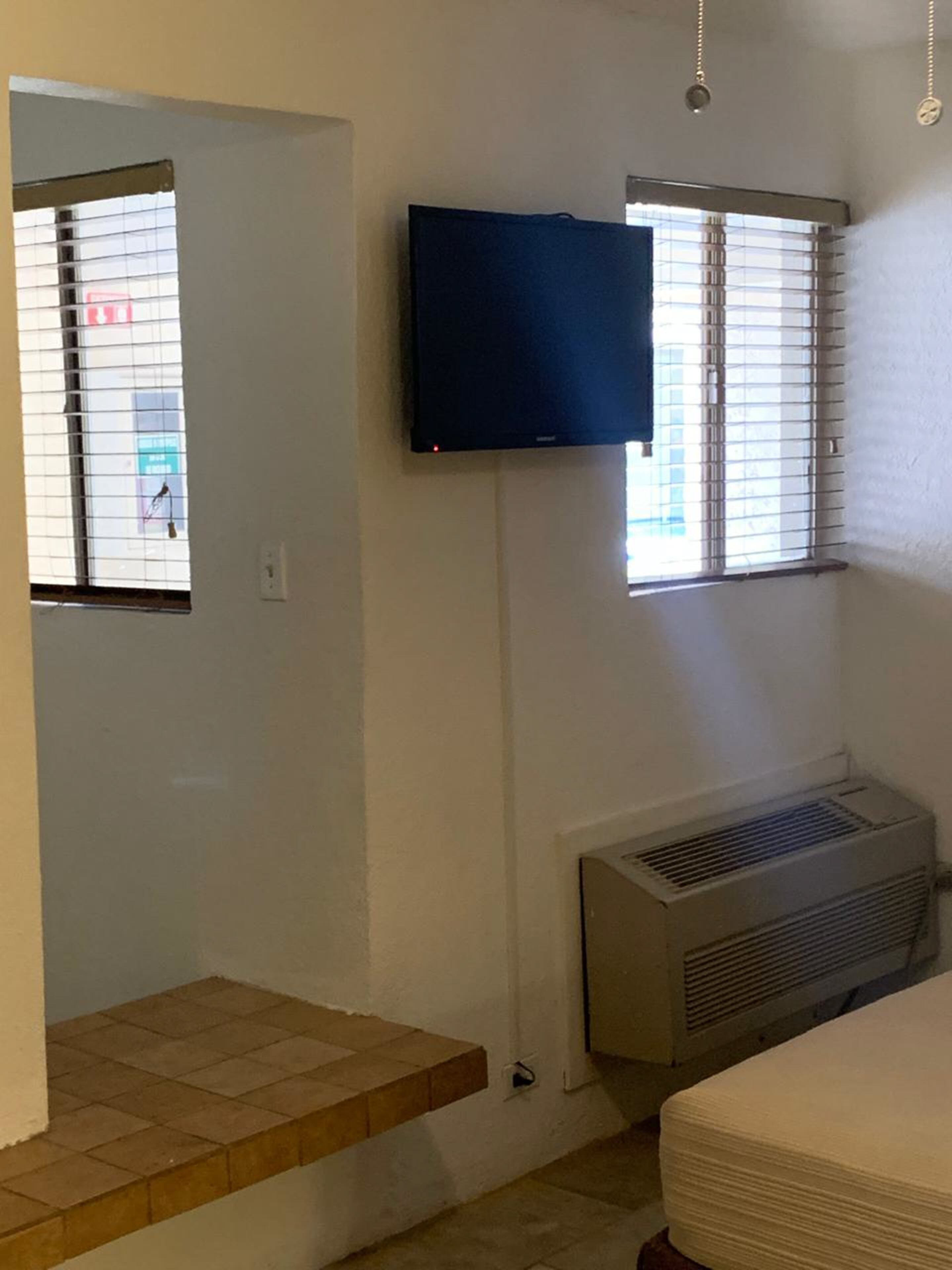 A room with a flat-screen TV mounted on the wall, a window with blinds, a radiator, and a small step leading to a door.