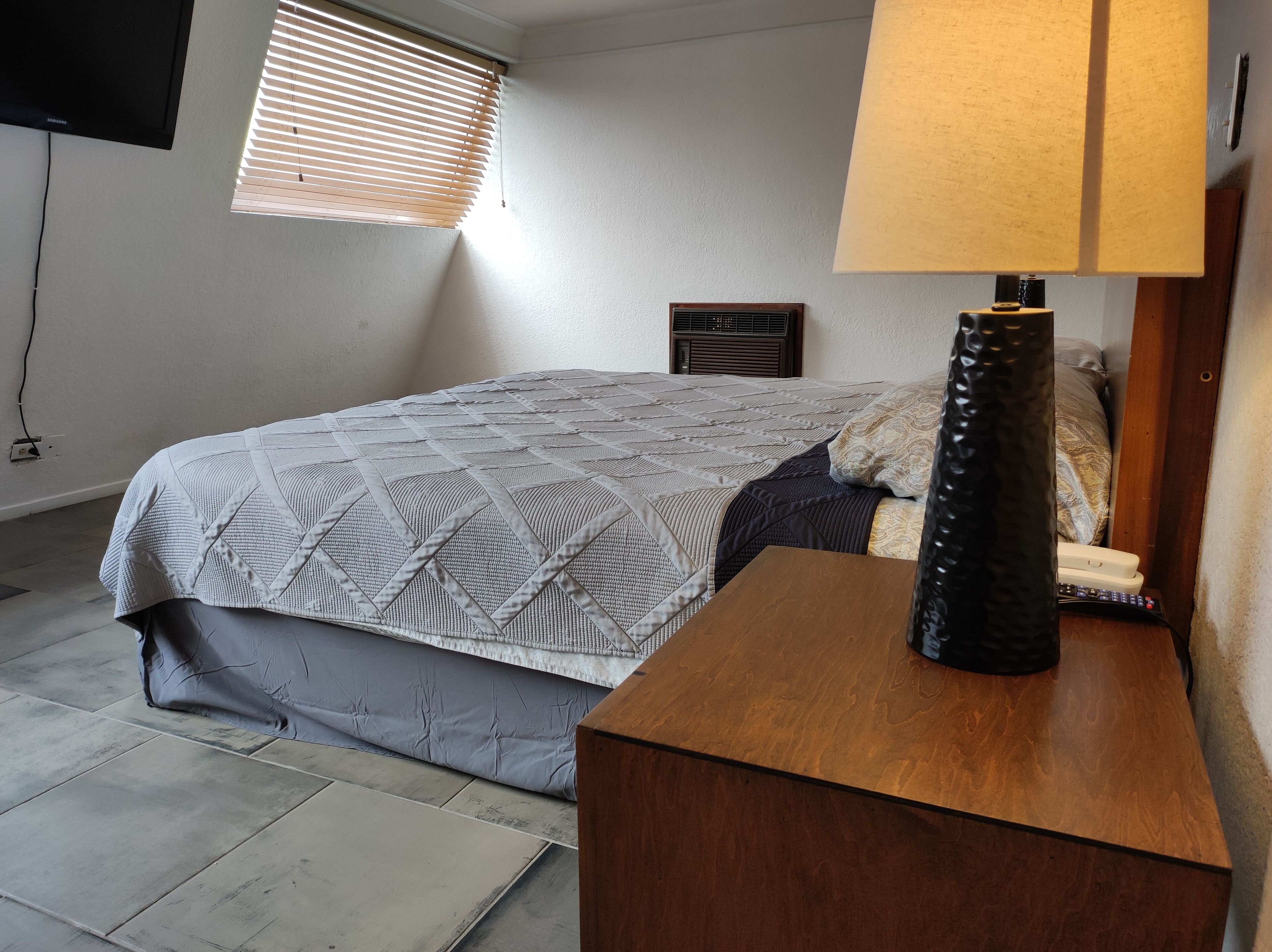 A bedroom with a bed, a bedside table, a lamp, a TV, and a window with blinds.