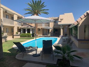 Un área de piscina con sillas de descanso y sombrillas, rodeada por un edificio con techo de tejas y una palmera.