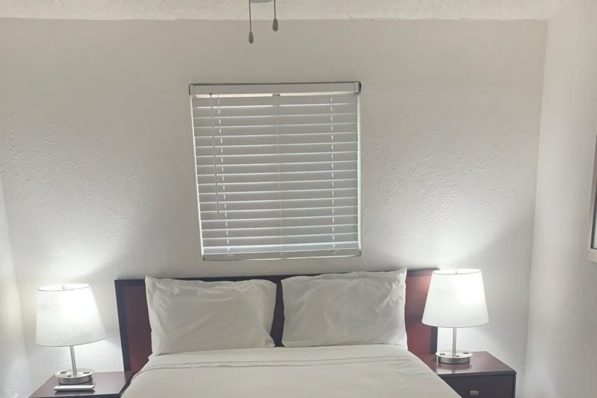 A neatly made bed with a striped skirt, two bedside tables with lamps, a window with blinds, and a ceiling fan.