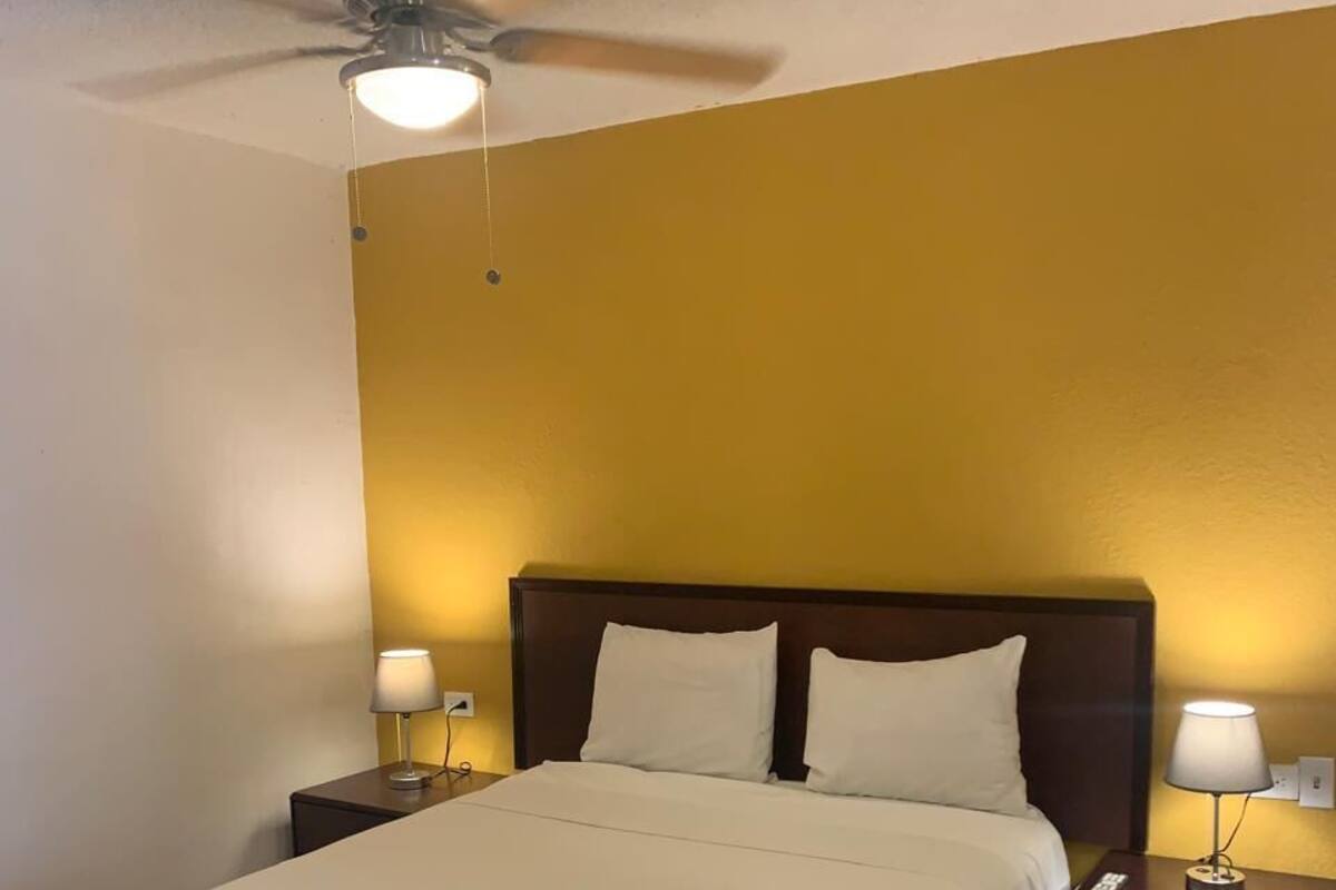 A hotel room with a bed, two bedside lamps, a ceiling fan, and a plain yellow wall.