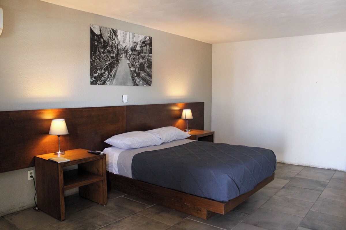 Un dormitorio ordenado con cabecera de madera, una cama individual con colcha azul oscuro, dos lámparas de noche y una fotografía blanco y negro en la pared.