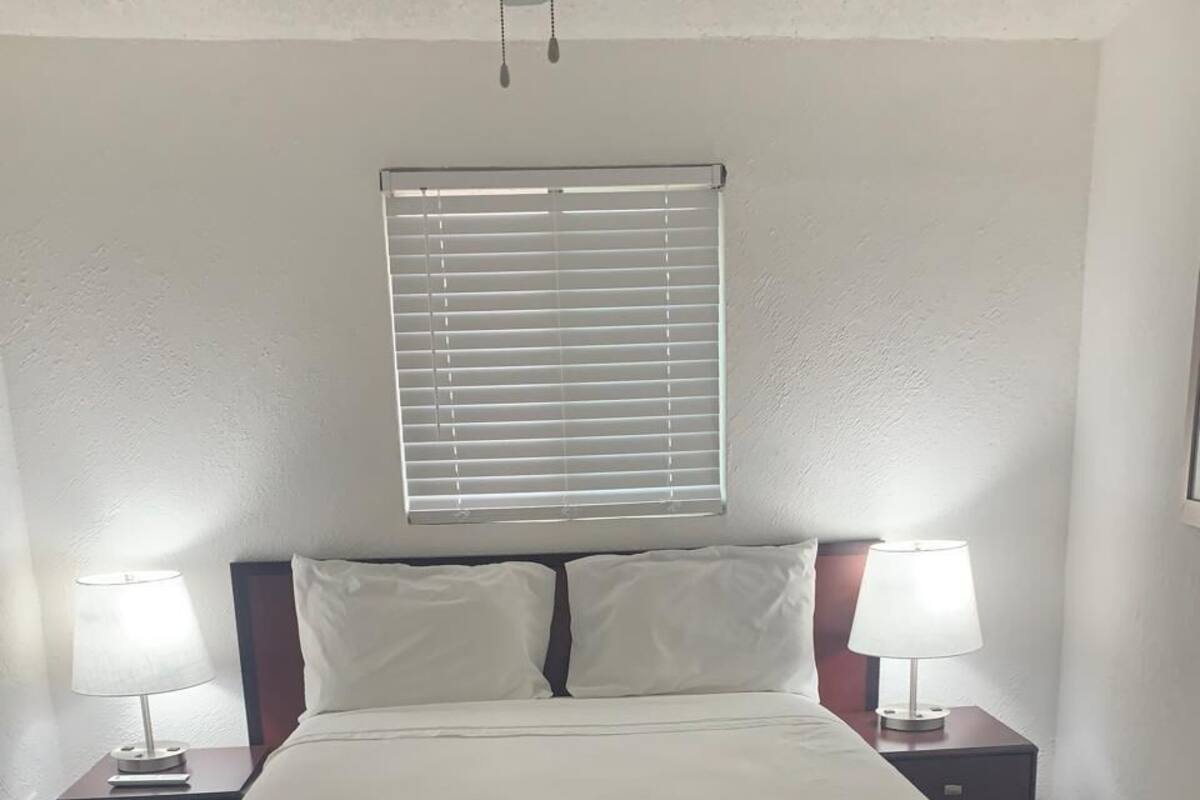 A neatly made bed with a striped skirt, two bedside tables with lamps, a window with blinds, and a ceiling fan.
