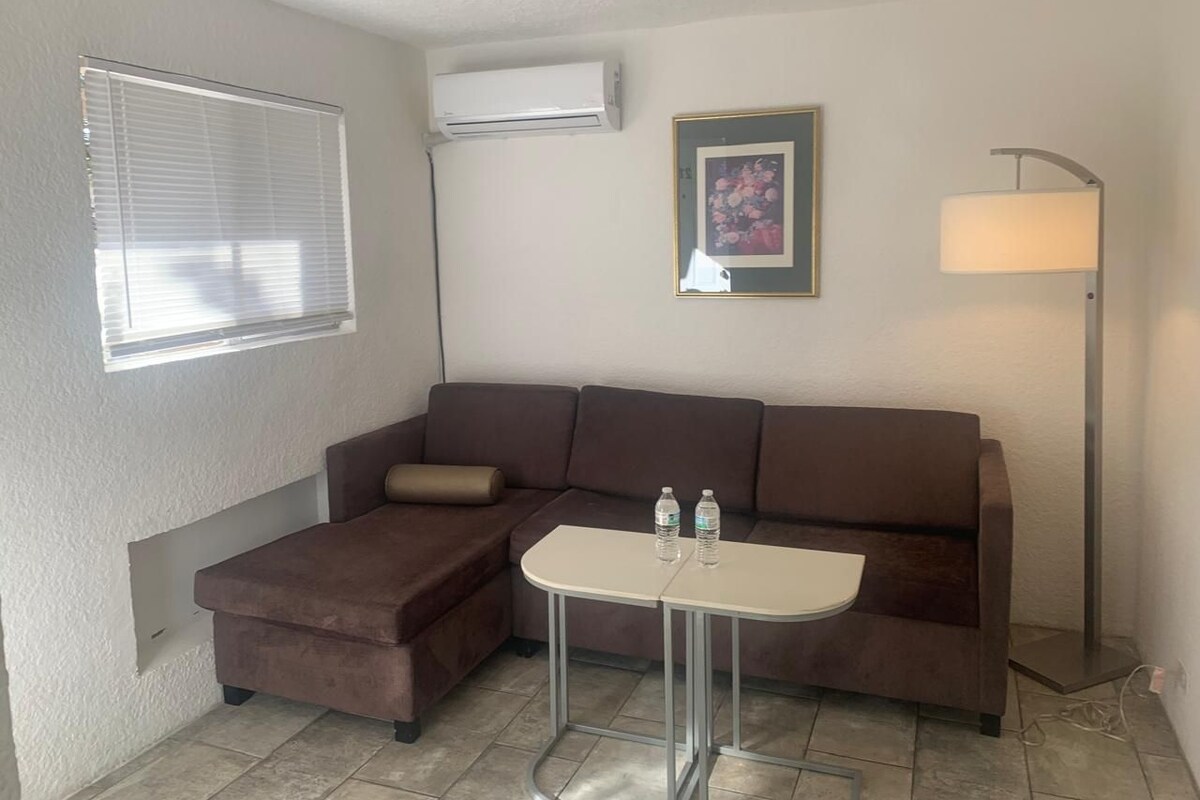 A small, furnished room with a brown sofa, a small table, a lamp, and an air conditioning unit on the wall.