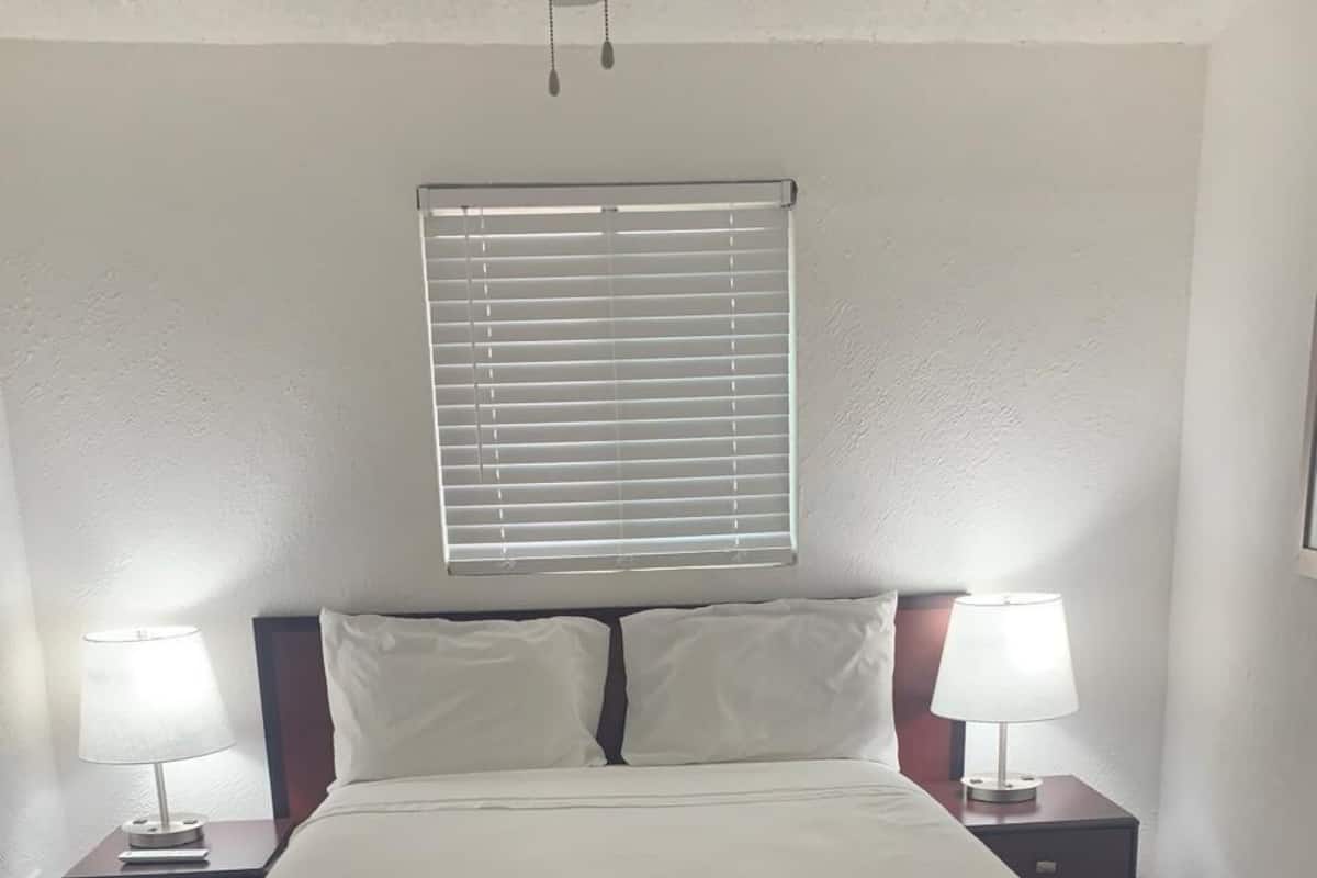 Una cama bien tendida con falda a rayas, dos mesitas de noche con lámparas, una ventana con persianas y un ventilador de techo.