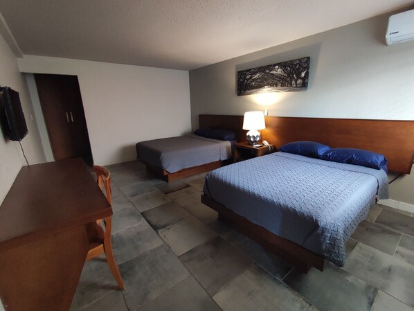 Habitación de hotel con dos camas, un escritorio y una silla. Hay un televisor de montaje en pared y un cuadro sobre las camas.