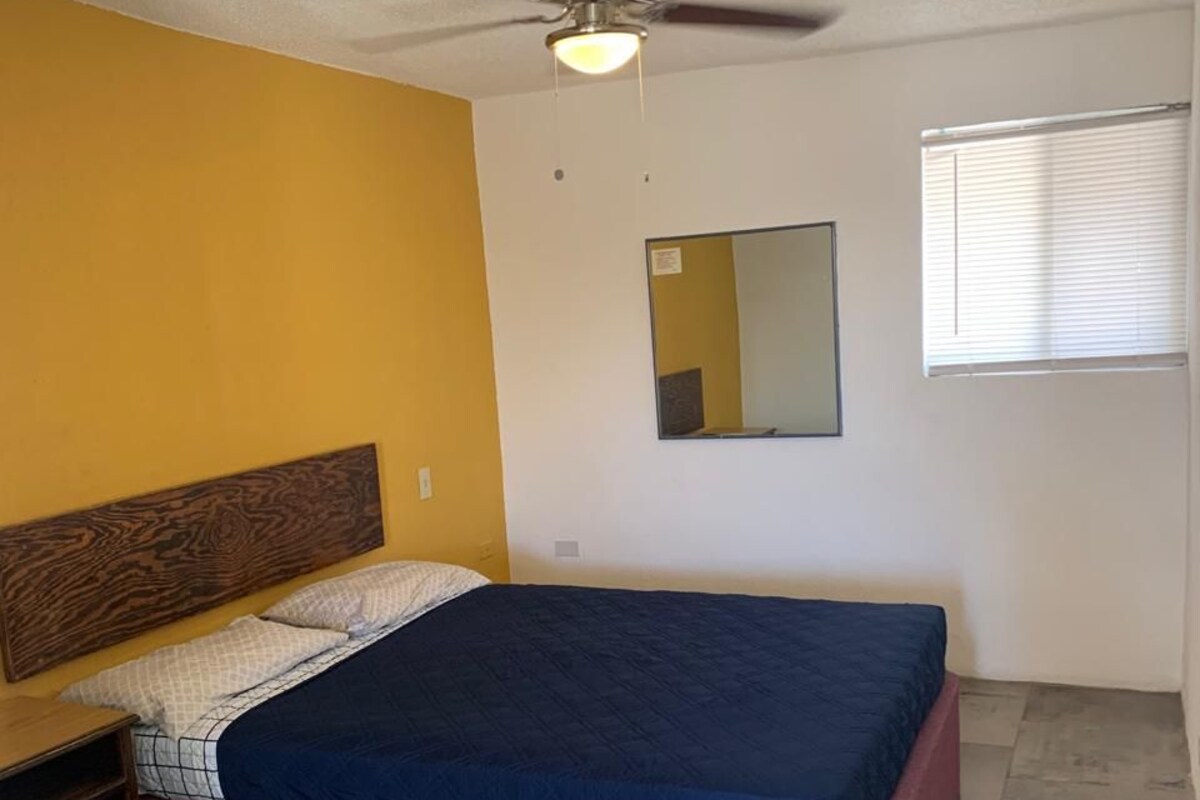 Un dormitorio con cama, ventilador de techo, mesita de noche, espejo y ventana.