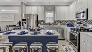 A modern kitchen with white cabinets, stainless steel appliances, a central island with stools, and a backsplash with a geometric pattern. - Modern Beach Retreat • Hot Tub • Private Beach Access • Sleeps 10 (Miramar Beach)