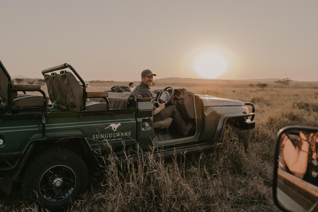 A safari vehicle with the brand name 'SUNGULWANE' and a logo, parked in a grassy field with a sunset in the background.