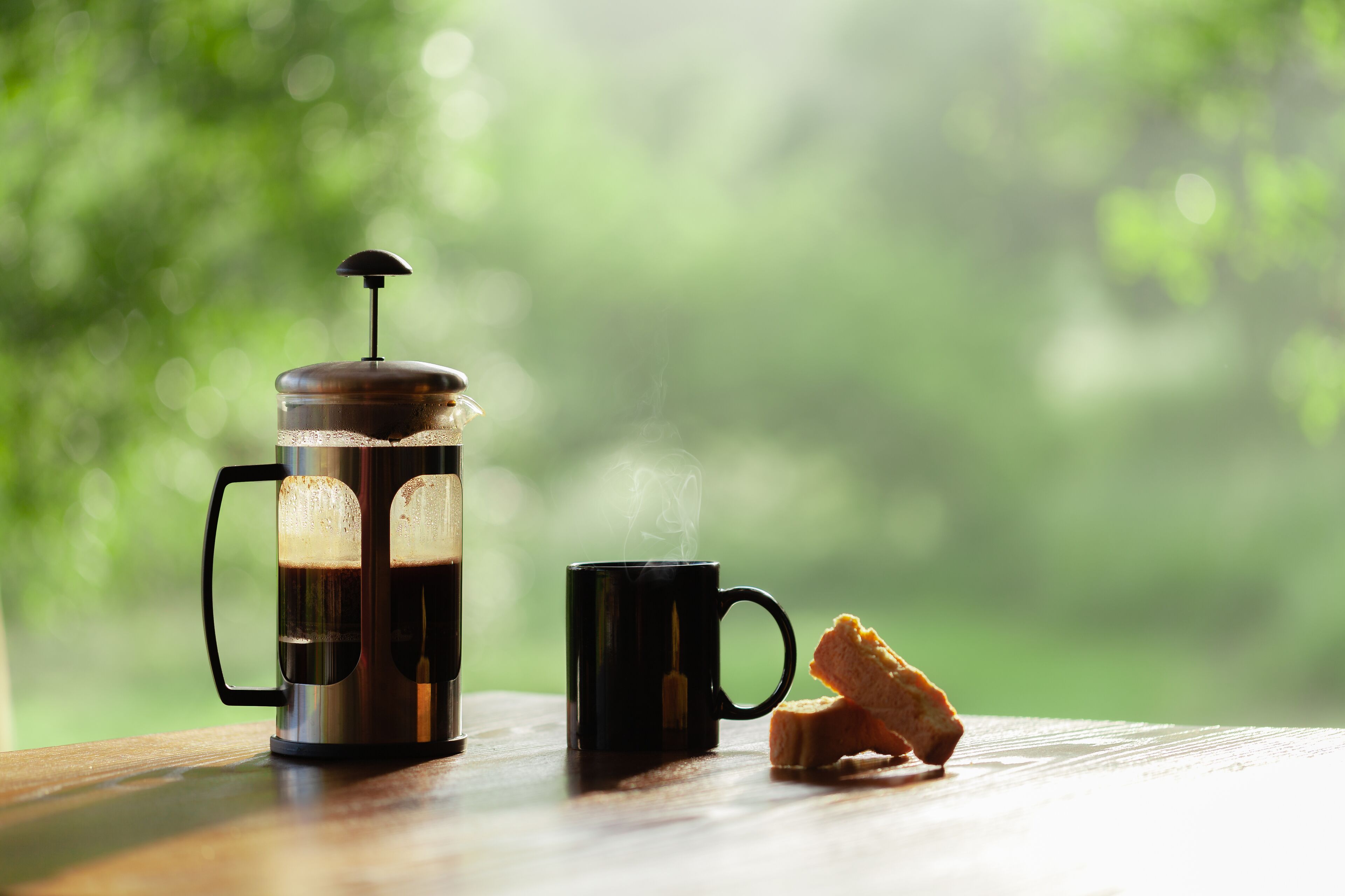A French press coffee maker with a lid and handle, a black mug, and a piece of bread on a wooden table with a blurred green background.
