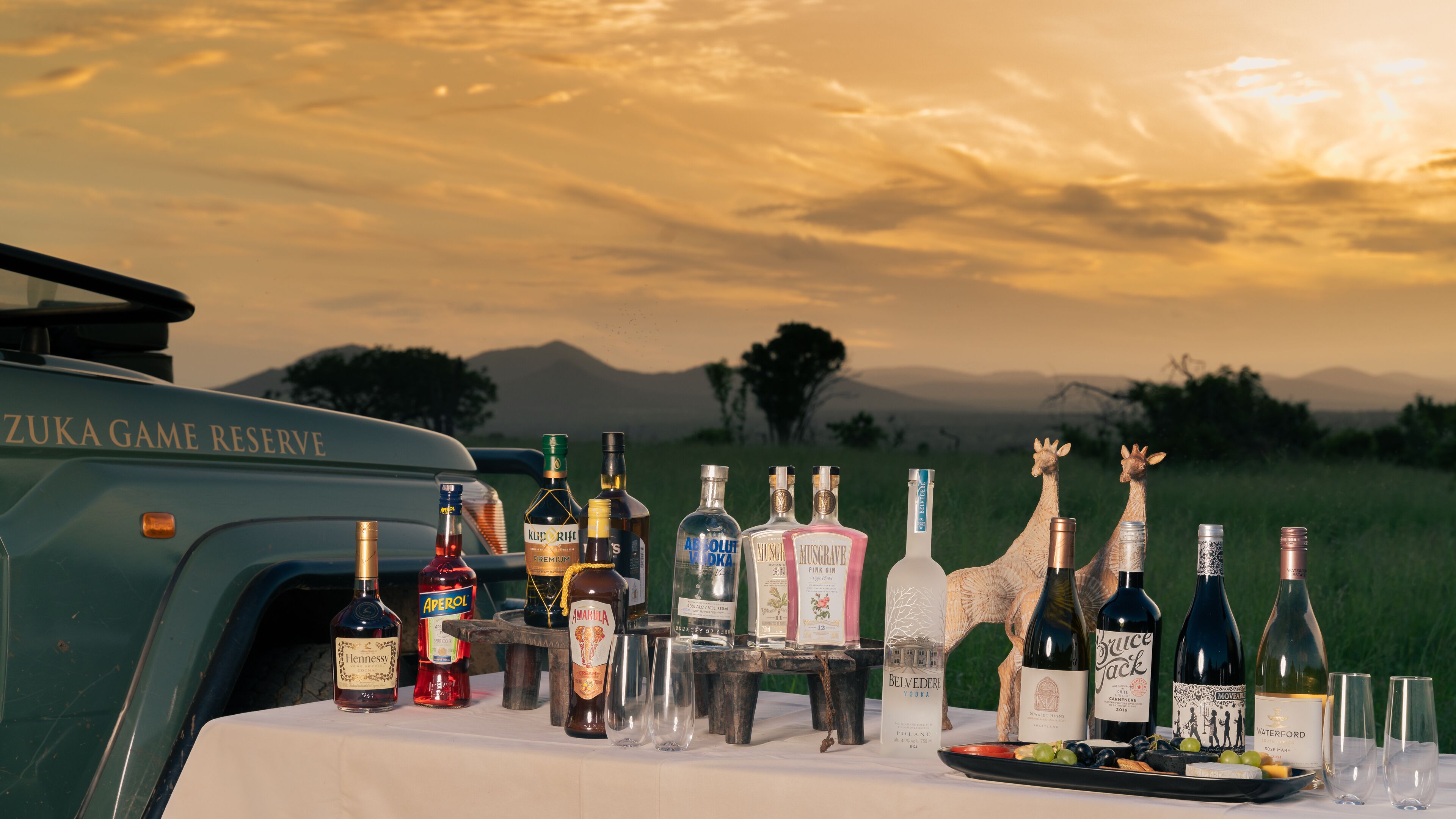 Una mesa con diversas botellas de alcohol y vasos, con un hermoso paisaje de atardecer y montañas al fondo.