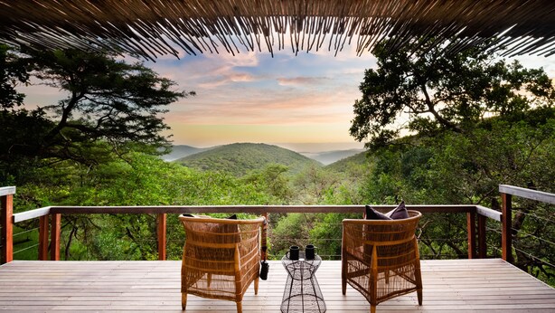 A wooden deck with wicker chairs and a metal table, overlooking a lush valley and mountains. - Sungulwane Private Game Lodge (Hluhluwe)