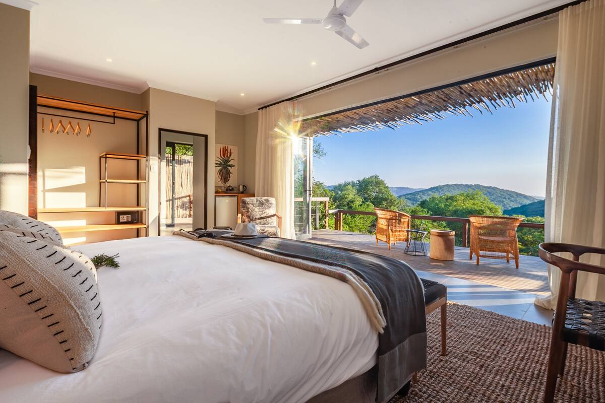A bedroom with a large bed, a ceiling fan, a view of the outdoors, and a balcony with seating.