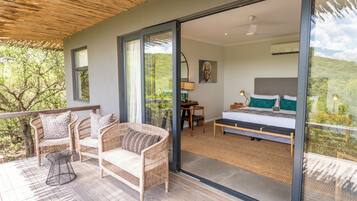 A bedroom with a bed, a desk, and a chair, a balcony with wicker furniture, and a view of the outdoors.